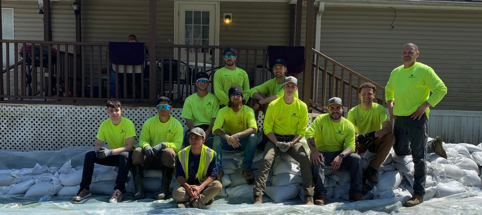 A group of ten Industrial Electricians in neon yellow shirts and one in a neon vest sit and stand on stacked sandbags in front of a porch, posing for a photo in daylight.
