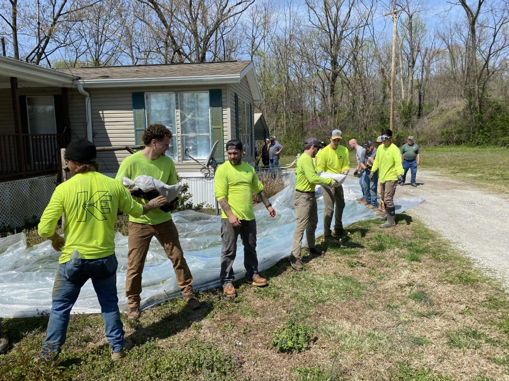 A group of people in yellow shirts, including industrial electricians, form a line outside a house, passing bags or materials along a plastic sheet on the ground. Trees and grass are visible in the background on a sunny day.