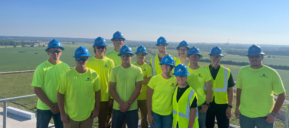 A group of Industrial Electricians in bright yellow shirts, safety vests, and blue hard hats stand together outdoors on a platform with a scenic green landscape and clear blue sky in the background.