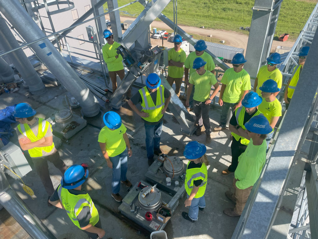 A group of construction workers in blue helmets and yellow safety vests gather around equipment on a metal platform at an industrial site, participating in a meeting or briefing.