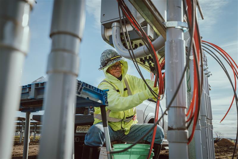 A construction worker in a hard hat and high-visibility jacket, like many Industrial Electricians, installs or repairs electrical wires on outdoor equipment at a construction site, surrounded by cables and metal structures.