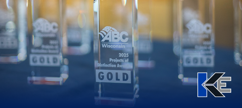 A close-up of a clear glass trophy labeled ABC Wisconsin 2025 Projects of Distinction Awards GOLD, honoring top Industrial Electrical Contractors, with similar trophies blurred in the background and a KE logo in the bottom right corner.