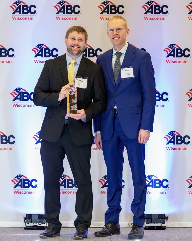 Two men in suits stand smiling in front of an ABC Wisconsin backdrop. One, holding a glass award, represents leading Industrial Electrical Contractors. Both wear nametags and pose proudly for a photo at the event.
