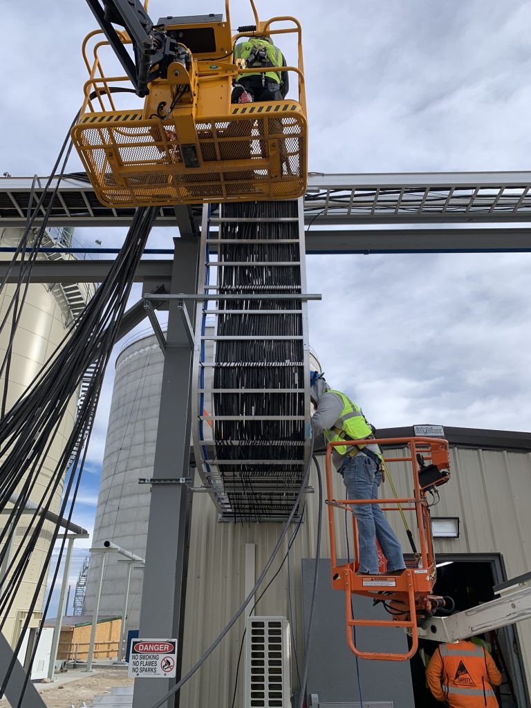 Two construction workers in safety gear operate orange lifts to access a large, vertical cable tray on an industrial site, where Industrial Electrical Contractors work near silos and a gray building in the background.