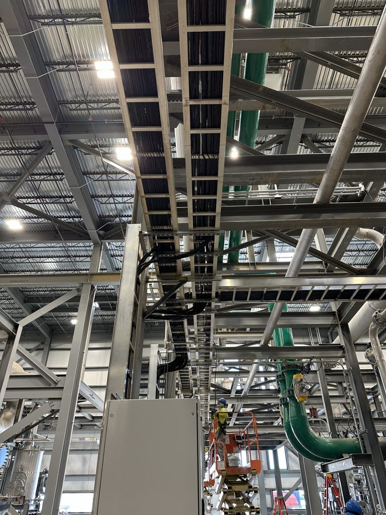 A view inside an industrial facility showing exposed metal beams, cable trays with wiring overhead, green pipes, and Industrial Electricians on a lift performing maintenance or installation work.