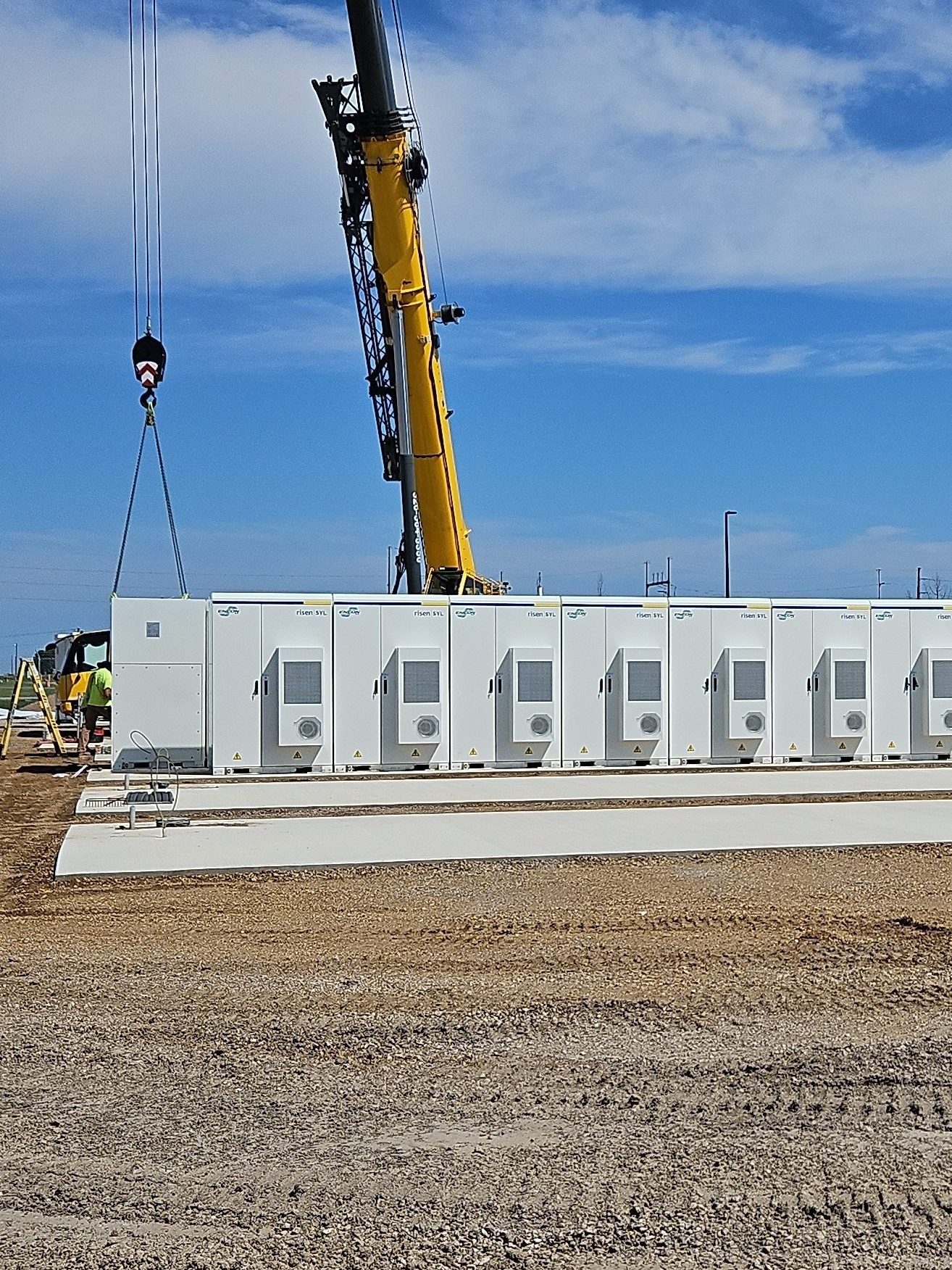 A crane lifts a large white industrial electrical unit onto a concrete pad as Industrial Electricians in safety gear stand nearby at the construction site, with blue sky and clouds in the background.