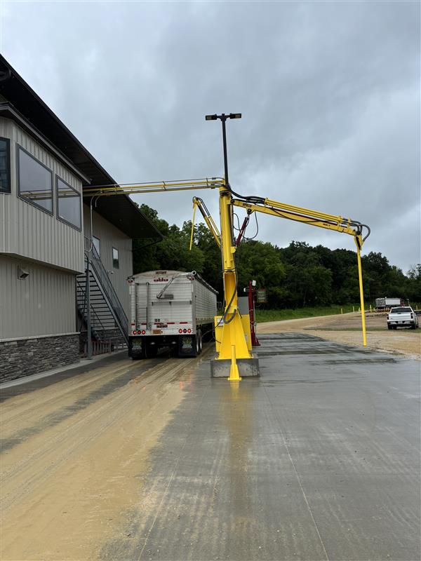 A yellow overhead loading arm, often installed by Industrial Electrical Contractors, extends from a building toward the back of a large transport truck parked on wet pavement, with trees and a white truck visible in the background under a cloudy sky.