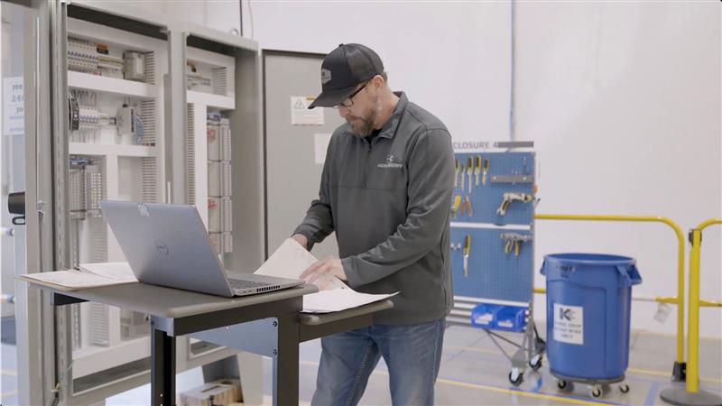 A man wearing a cap and glasses stands at a desk with a laptop and papers in an industrial setting, with an open control panel, a blue trash bin, and a tool board in the background.