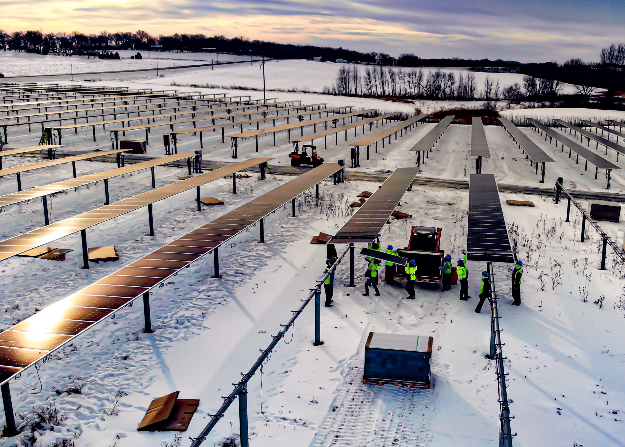 Workers in safety vests install solar panels on large metal frames in a snowy field, with multiple rows of solar panels stretching into the distance under a cloudy sky.