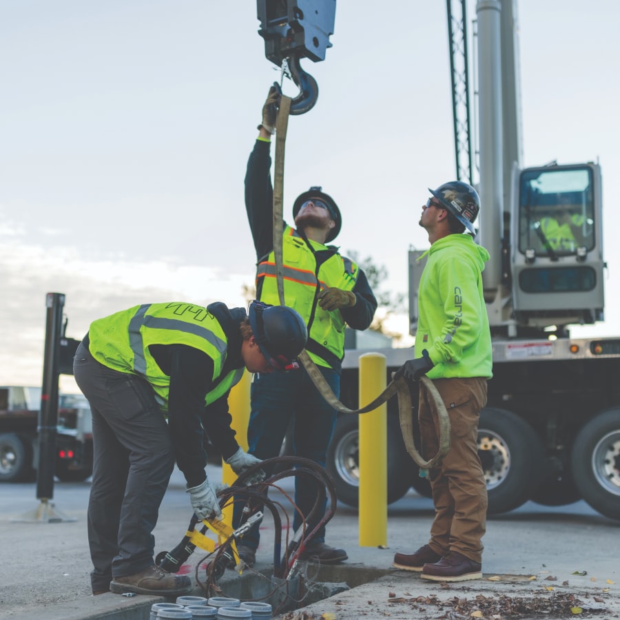 Three construction workers wearing safety vests and helmets operate heavy machinery outdoors; one attaches a hook to equipment, while another bends down working with cables and tools.