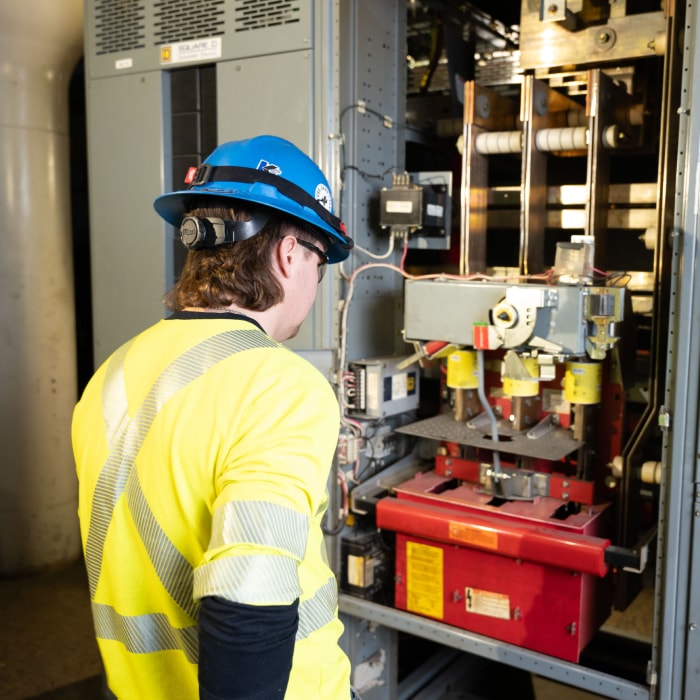 A worker in a yellow safety shirt and blue hard hat inspects an open electrical panel with various switches and components in an industrial setting.