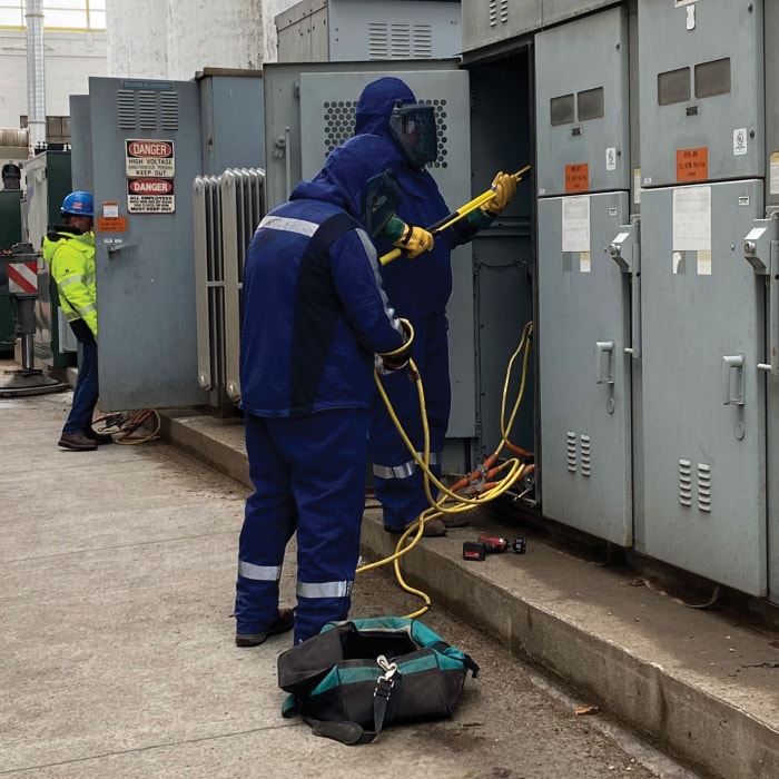 Two workers in blue protective suits and helmets handle electrical equipment at an outdoor industrial site, inspecting large gray electrical panels. Another person in a safety vest stands nearby. Tools and a bag are on the ground.