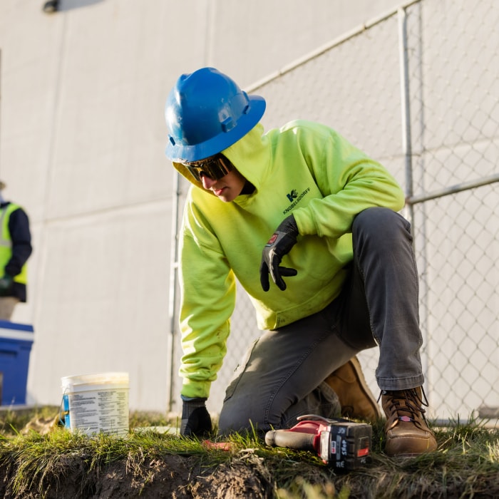 A construction worker in a neon yellow hoodie, blue hard hat, and gloves kneels on the grass by a fence, working with tools and a plastic container on the ground.