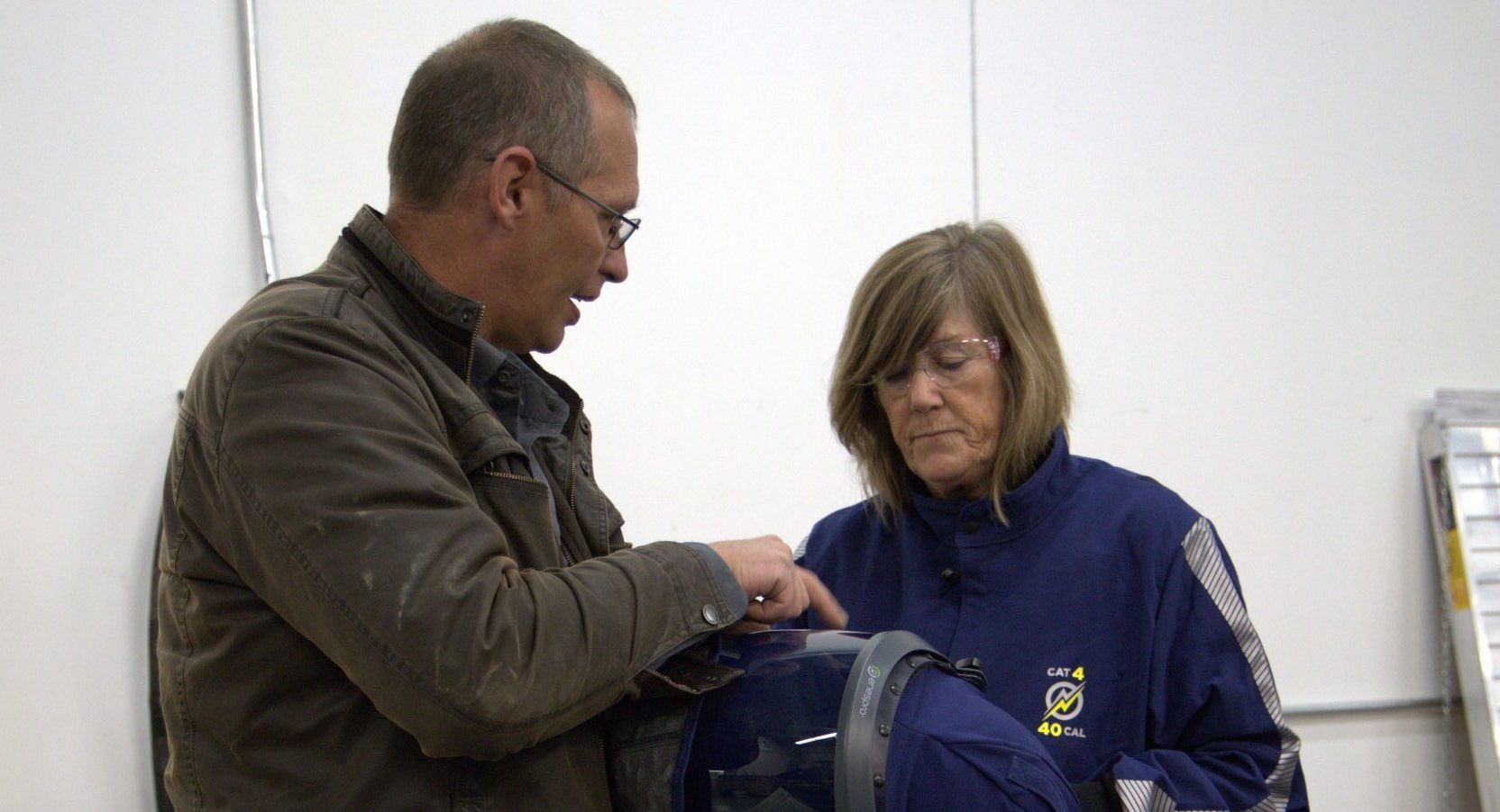 A man helps a woman put on safety gear in an indoor setting. The woman is wearing a blue uniform and the man is wearing a brown jacket. Various equipment and bags are visible around them.
