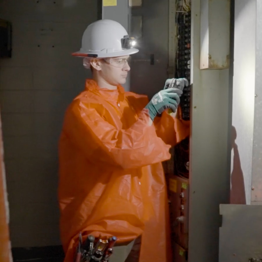 A worker wearing an orange protective suit, hard hat with headlamp, gloves, and safety glasses inspects an electrical panel with a tool, surrounded by various instruments.