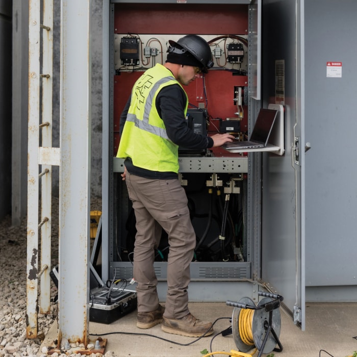 A worker wearing a hard hat and safety vest uses a laptop at an open electrical control panel outdoors. Tools and a yellow extension cord are on the ground nearby.