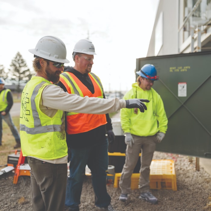 Three construction workers wearing safety vests, hard hats, and gloves stand outdoors. One worker in a yellow vest points ahead while the others listen attentively near a large green utility box.