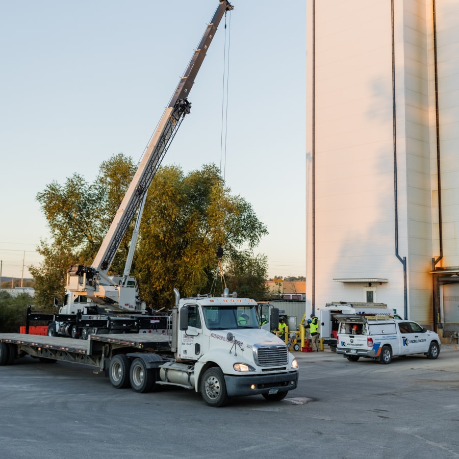 A crane lifts equipment from a flatbed truck near a tall industrial building, with workers and utility vehicles nearby, in the early morning or late afternoon light.