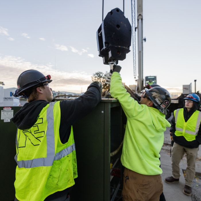 Two construction workers in safety gear and helmets guide a large hook attached to a crane, while a third worker stands nearby on a building site during daylight.