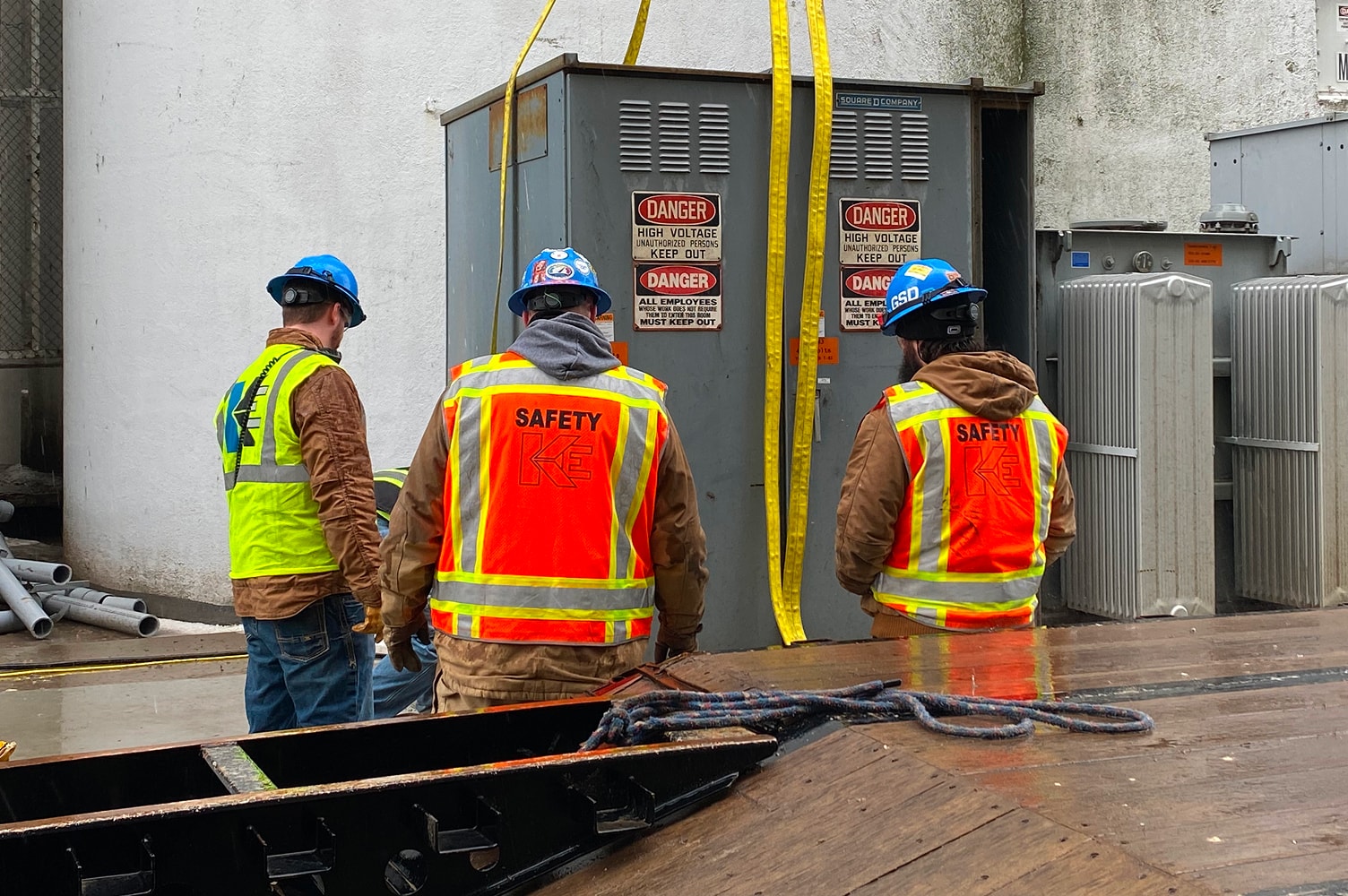 Three construction workers wearing safety vests and helmets stand near a large electrical unit with “Danger High Voltage” signs, discussing the job site.
