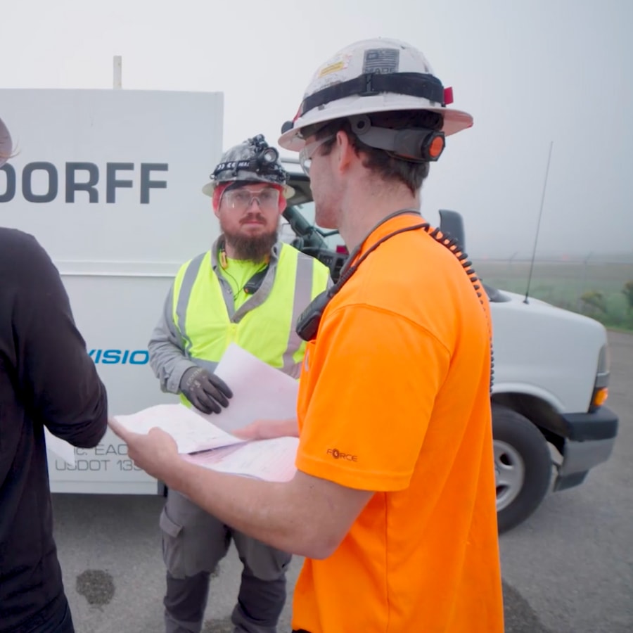 Three construction workers, two in safety gear and one in a bright orange shirt and hard hat, stand near a white van discussing papers on a foggy day at a worksite.