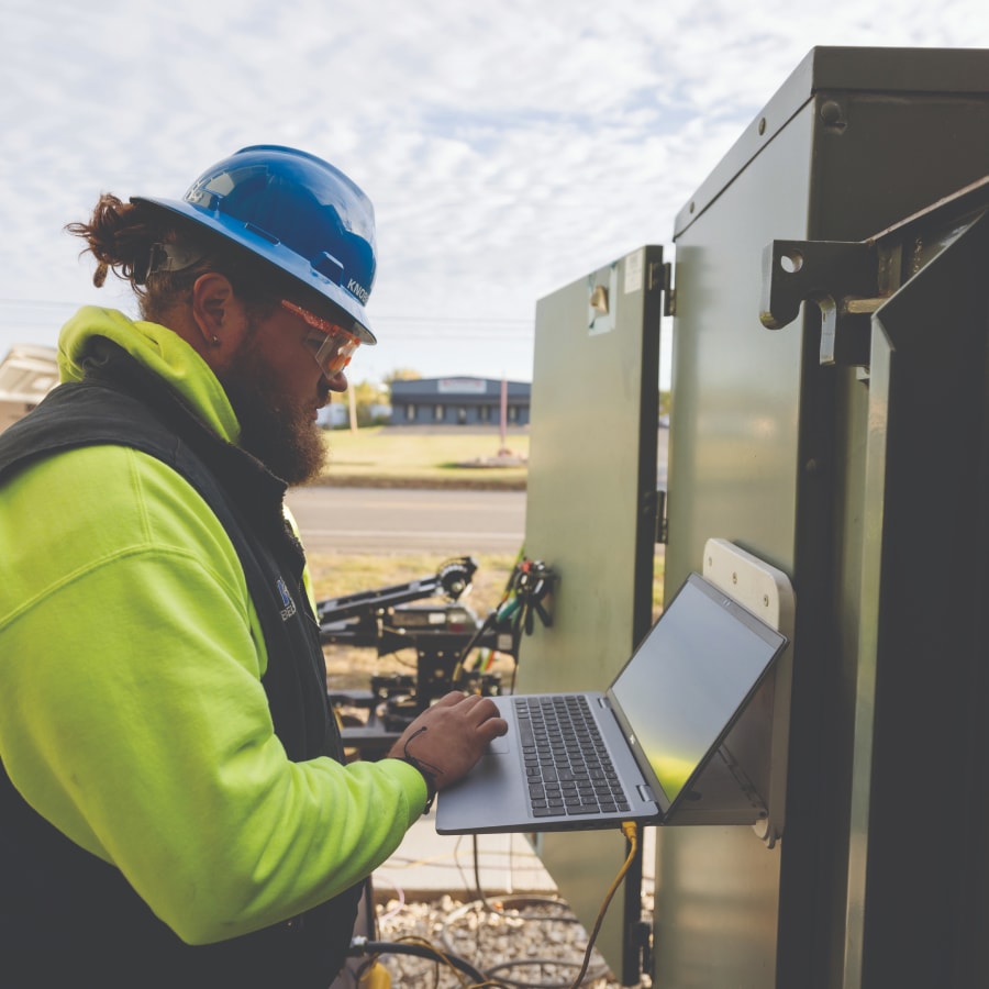 A worker in a blue hard hat and neon yellow jacket uses a laptop mounted on an open outdoor electrical cabinet near a road, with equipment and buildings in the background.